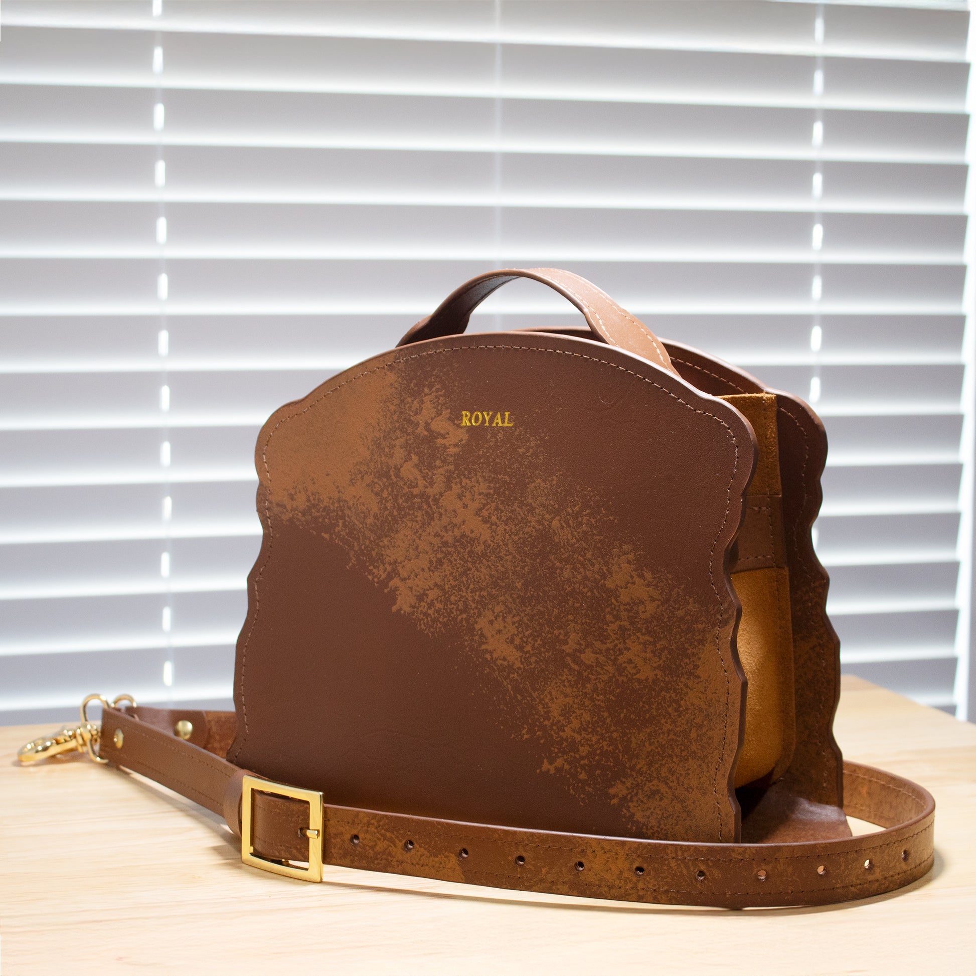 Hand‑painted full‑grain leather Joy satchel handcrafted in brown with artisan multi‑tone accents, shown on a light wooden surface with gold‑tone hardware.  